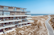 A modern multi-story building with sleek glass balconies overlooks a sandy beach with scattered vegetation. A road winds along the edge of the beach, leading to the horizon. The ocean is calm and the sky is clear and blue.