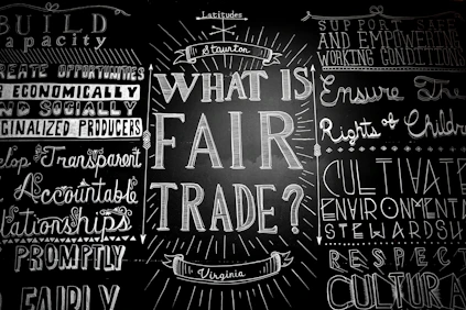 A blackboard with various words and phrases related to fair trade, written in decorative fonts. The central question 'What is Fair Trade?' is prominently featured, surrounded by other concepts like 'support safe working conditions' and 'develop transparent relationships.'