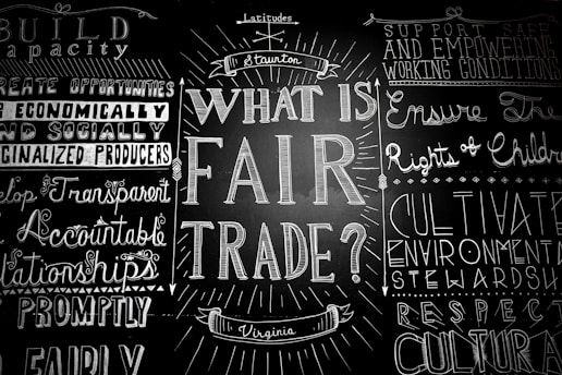 A blackboard with various words and phrases related to fair trade, written in decorative fonts. The central question 'What is Fair Trade?' is prominently featured, surrounded by other concepts like 'support safe working conditions' and 'develop transparent relationships.'