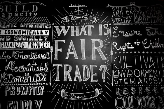 A blackboard with various words and phrases related to fair trade, written in decorative fonts. The central question 'What is Fair Trade?' is prominently featured, surrounded by other concepts like 'support safe working conditions' and 'develop transparent relationships.'