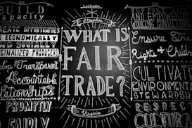 A blackboard with various words and phrases related to fair trade, written in decorative fonts. The central question 'What is Fair Trade?' is prominently featured, surrounded by other concepts like 'support safe working conditions' and 'develop transparent relationships.'