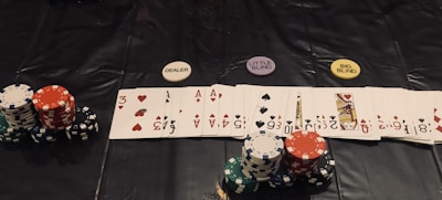 A poker game setup is displayed, featuring stacks of colorful poker chips including red, green, black, and white. Playing cards are laid out in a row, revealing various suits and numbers. Three distinct dealer buttons labeled 'DEALER', 'LITTLE BLIND', and 'BIG BLIND' are positioned above the card lineup.