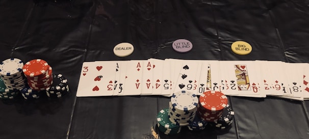 A poker game setup is displayed, featuring stacks of colorful poker chips including red, green, black, and white. Playing cards are laid out in a row, revealing various suits and numbers. Three distinct dealer buttons labeled 'DEALER', 'LITTLE BLIND', and 'BIG BLIND' are positioned above the card lineup.