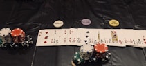 A poker game setup is displayed, featuring stacks of colorful poker chips including red, green, black, and white. Playing cards are laid out in a row, revealing various suits and numbers. Three distinct dealer buttons labeled 'DEALER', 'LITTLE BLIND', and 'BIG BLIND' are positioned above the card lineup.