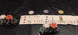 A poker game setup is displayed, featuring stacks of colorful poker chips including red, green, black, and white. Playing cards are laid out in a row, revealing various suits and numbers. Three distinct dealer buttons labeled 'DEALER', 'LITTLE BLIND', and 'BIG BLIND' are positioned above the card lineup.