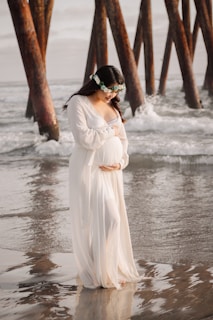Expectant mother wrapped in a soft shawl, standing on a sandy beach with waves in the background