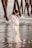 A pregnant woman in a flowing white dress stands on a sandy beach with the ocean in the background. She is gently holding her belly and wearing a floral headband. The scene includes wooden pillars from a pier and waves softly crashing against the shore.