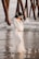 A pregnant woman in a flowing white dress stands on a sandy beach with the ocean in the background. She is gently holding her belly and wearing a floral headband. The scene includes wooden pillars from a pier and waves softly crashing against the shore.