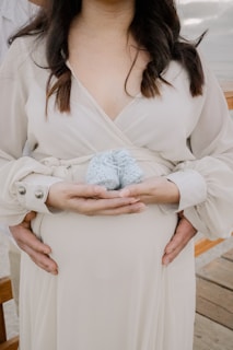 Hands gently cradle a pregnant belly, with a small pair of baby shoes held in the center. The wearer is dressed in a flowing cream dress and stands in a serene outdoor setting.