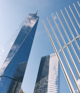 Tall modern skyscrapers with reflective glass exteriors reaching into a clear blue sky. Vertical white structures are in the foreground, adding a dynamic architectural element.
