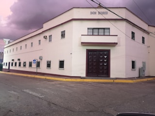 A rectangular, two-story building with a sign that reads 'Don Bosco'. It has multiple windows with security bars and a large, dark wooden door at the front. The building is painted white with maroon trim, and there are utility wires crossing in front. The sky appears overcast with purple hues, suggesting a potential storm. The building is situated on a paved street with a curb.