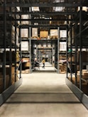 Long aisles filled with metal shelves holding boxes and packages in a warehouse setting. A person in a yellow shirt is pushing a cart down the aisle, with rows of organized shelves extending into the background.