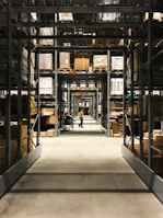 Long aisles filled with metal shelves holding boxes and packages in a warehouse setting. A person in a yellow shirt is pushing a cart down the aisle, with rows of organized shelves extending into the background.