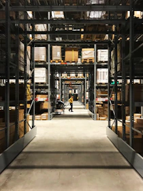 Long aisles filled with metal shelves holding boxes and packages in a warehouse setting. A person in a yellow shirt is pushing a cart down the aisle, with rows of organized shelves extending into the background.