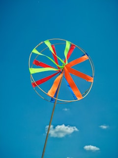 A colorful wind spinner with vibrant ribbons of orange, green, red, and blue attached to a circular frame, set against a bright blue sky. A few small white clouds are scattered in the background, creating a cheerful and playful ambiance.