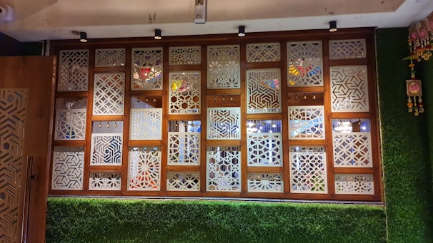 A decorative wall panel featuring wooden frames with intricate geometric lattice patterns. The panel includes small hanging bells and is surrounded by artificial grass on the lower section and sides. Multicolored glass elements on the other side can be seen through the patterns.