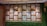 A decorative wall panel featuring wooden frames with intricate geometric lattice patterns. The panel includes small hanging bells and is surrounded by artificial grass on the lower section and sides. Multicolored glass elements on the other side can be seen through the patterns.