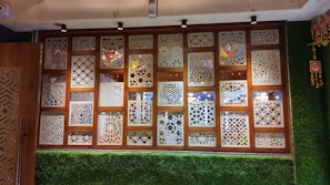 A decorative wall panel featuring wooden frames with intricate geometric lattice patterns. The panel includes small hanging bells and is surrounded by artificial grass on the lower section and sides. Multicolored glass elements on the other side can be seen through the patterns.