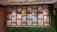 A decorative wall panel featuring wooden frames with intricate geometric lattice patterns. The panel includes small hanging bells and is surrounded by artificial grass on the lower section and sides. Multicolored glass elements on the other side can be seen through the patterns.
