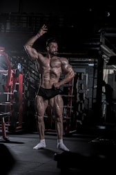 A muscular man poses confidently in a dimly lit gym, highlighting his well-defined physique. He is wearing minimal clothing to showcase his musculature, with gym equipment such as dumbbells and a squat rack in the background.