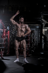 A muscular man poses confidently in a dimly lit gym, highlighting his well-defined physique. He is wearing minimal clothing to showcase his musculature, with gym equipment such as dumbbells and a squat rack in the background.