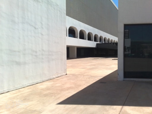 A modern architectural structure with a white facade and a series of evenly spaced arches along the second level. The ground is paved with light-colored concrete, casting sharp shadows in the bright sunlight. A large wall in the foreground creates a division and there is a dark glass window on the right side.