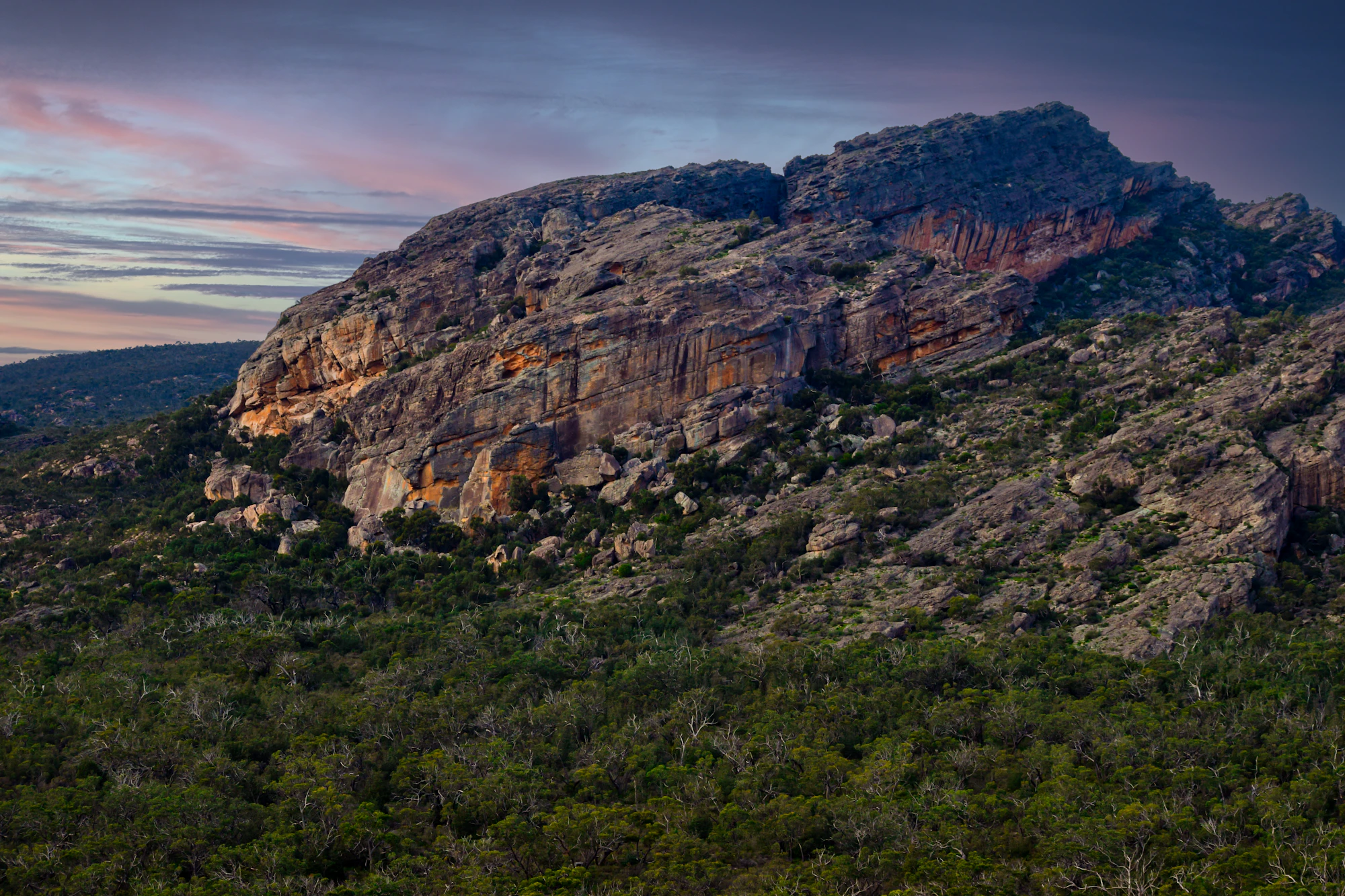 A rugged sandstone rock formation rising above dense woodland beneath a colourful evening sky.