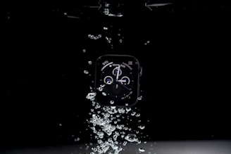A smartwatch submerged in water with visible air bubbles surrounding it, the watch face displays various dials and indicators.