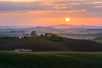 A picturesque landscape features rolling green hills under a softly glowing sunset. In the mid-ground, a traditional countryside estate with several buildings is surrounded by trees. The sky is a gentle gradient of orange and pink hues, and the sun hangs low, casting warm light over the scene.