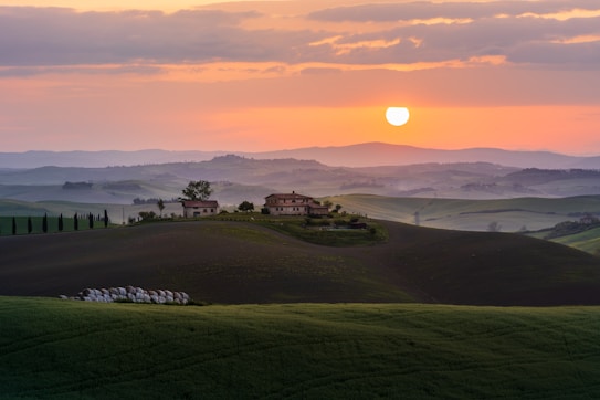 A picturesque landscape features rolling green hills under a softly glowing sunset. In the mid-ground, a traditional countryside estate with several buildings is surrounded by trees. The sky is a gentle gradient of orange and pink hues, and the sun hangs low, casting warm light over the scene.