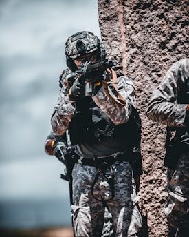 A soldier in camouflage gear is aiming a rifle while standing next to a textured wall. The environment appears rugged and outdoor.