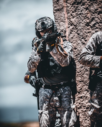 An action photo of an airsoft player in camo gear aiming a replica rifle in a wooded area.