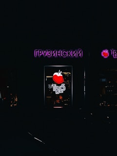 A dimly lit storefront with neon signage featuring text in Cyrillic script. The central focus is a neon red apple with the letters 'TBLS' below it in bright white. There is a faint warm light from the interior, hinting at a cozy atmosphere inside.