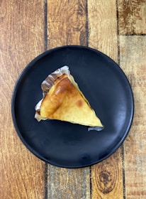 A triangular slice of cake on a black plate, placed on a wooden surface. The cake is wrapped partially in silver foil, with a golden-brown crust and a smooth, creamy top.