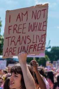 A protest scene with a person holding up a cardboard sign with the words 'I AM NOT FREE WHILE TRANS LIVES ARE UNFREE' written in bold red letters. The setting is outdoors on a sunny day, with a crowd of people in the background, some holding similar signs. The mood appears to be passionate and determined.