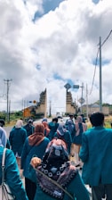 A group of people wearing teal jackets, some with headscarves, are walking along a road toward a traditional stone gateway. The sky is filled with large, fluffy clouds, and there are electrical poles and road signs visible in the background.