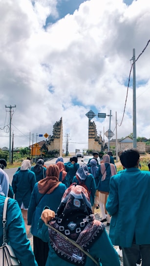 A group of people wearing teal jackets, some with headscarves, are walking along a road toward a traditional stone gateway. The sky is filled with large, fluffy clouds, and there are electrical poles and road signs visible in the background.