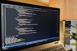 A laptop is displaying a code editor with HTML code on the screen. The environment is softly illuminated by a lamp with a brown shade, and there is a green plant in the background, contributing to a cozy workspace setting.