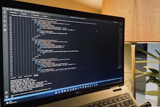 A laptop is displaying a code editor with HTML code on the screen. The environment is softly illuminated by a lamp with a brown shade, and there is a green plant in the background, contributing to a cozy workspace setting.