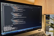 A laptop is displaying a code editor with HTML code on the screen. The environment is softly illuminated by a lamp with a brown shade, and there is a green plant in the background, contributing to a cozy workspace setting.