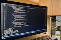 A laptop is displaying a code editor with HTML code on the screen. The environment is softly illuminated by a lamp with a brown shade, and there is a green plant in the background, contributing to a cozy workspace setting.
