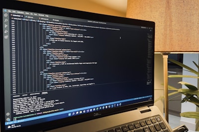 A laptop is displaying a code editor with HTML code on the screen. The environment is softly illuminated by a lamp with a brown shade, and there is a green plant in the background, contributing to a cozy workspace setting.