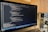 A laptop is displaying a code editor with HTML code on the screen. The environment is softly illuminated by a lamp with a brown shade, and there is a green plant in the background, contributing to a cozy workspace setting.