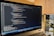 A laptop is displaying a code editor with HTML code on the screen. The environment is softly illuminated by a lamp with a brown shade, and there is a green plant in the background, contributing to a cozy workspace setting.