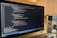 A laptop is displaying a code editor with HTML code on the screen. The environment is softly illuminated by a lamp with a brown shade, and there is a green plant in the background, contributing to a cozy workspace setting.