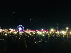 A large crowd at a nighttime outdoor event with many people holding up phones or lights. In the background, a brightly lit Ferris wheel and colorful flags can be seen, creating a festive atmosphere.