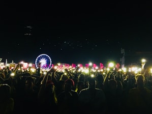 A large crowd at a nighttime outdoor event with many people holding up phones or lights. In the background, a brightly lit Ferris wheel and colorful flags can be seen, creating a festive atmosphere.