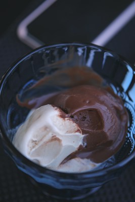 A glass dish containing two scoops of ice cream, one vanilla and the other chocolate, with a smooth and creamy texture. The background is dark, providing contrast to the lighter colors of the ice cream.