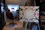 An intricately designed mandala on an easel dominates the foreground, featuring a symmetric pattern with vibrant colors like pink, yellow, and black. In the background, a workspace is visible, with a computer screen displaying an image or video, surrounded by various items such as speakers, a mannequin bust, and miscellaneous objects on a desk. A rolling chair with a red garment draped over it adds to the lived-in feel of the room.