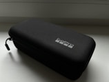 A black, rectangular case with a visible zipper and a small, branded logo on the top. The material appears to be textured, possibly fabric or hard-shell. The lighting casts a subtle shadow, suggesting it is placed on a flat surface near a light source.